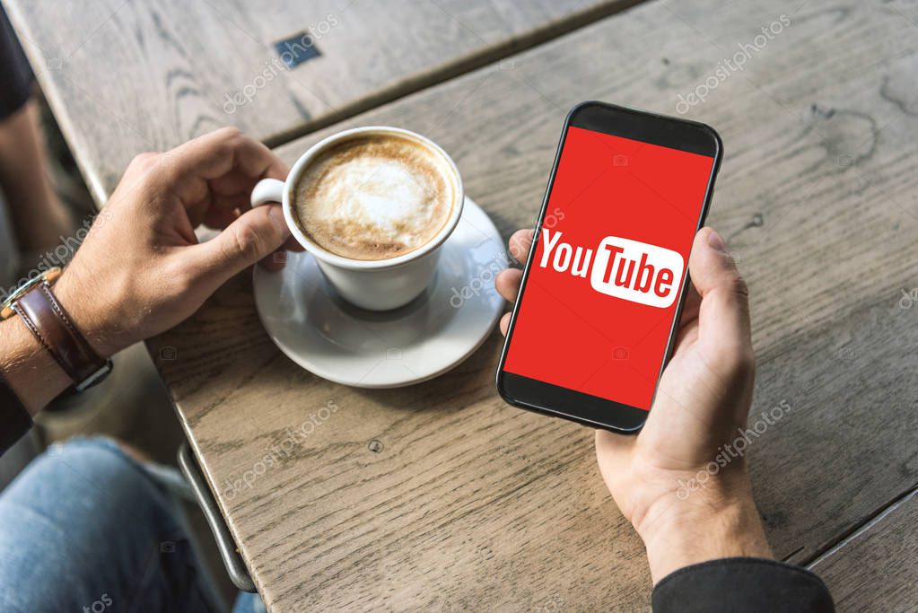 Cropped shot of man with cup of cappuccino using smartphone with youtube logo on screen