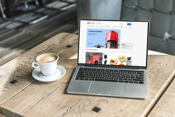 чашка кофе и ноутбук с веб-сайтом ebay на экране на деревенском деревянном столе в кафе
