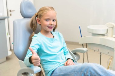 beautiful little child sitting in dental chair at dentist office and showing thumb up clipart