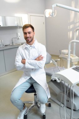handsome young dentist with crossed arms looking at camera while sitting at office clipart