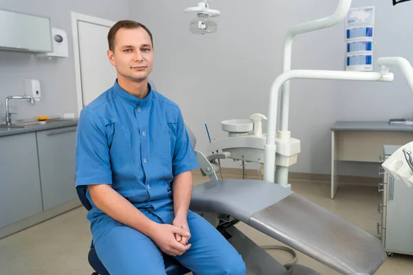 Przystojny Młody Dentysta Siedzi Gabinecie Stomatologicznym Patrząc Kamery — Zdjęcie stockowe