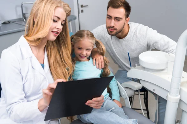 Hermosa Dentista Femenina Mostrando Portapapeles Con Diagnóstico Padre Hija Pequeña —  Fotos de Stock