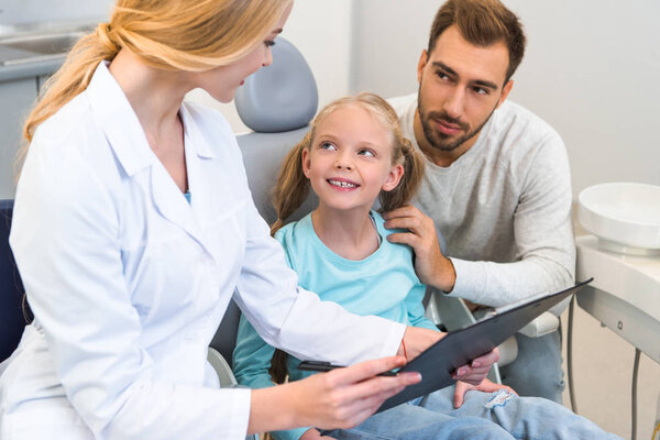 young female dentist showing clipboard with diagnosis to father and little daughter