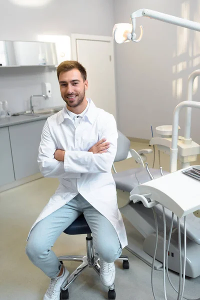 Bello Giovane Dentista Con Braccia Incrociate Guardando Fotocamera Mentre Seduto — Foto Stock