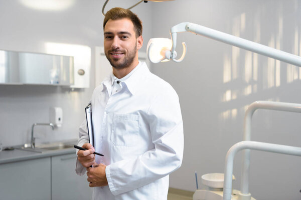 handsome young dentist in white coat with clipboard looking at camera in office