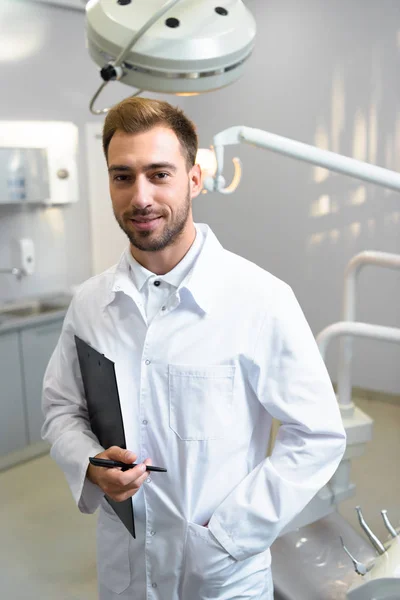 Smiling Young Dentist White Coat Clipboard Looking Camera Office — Free Stock Photo