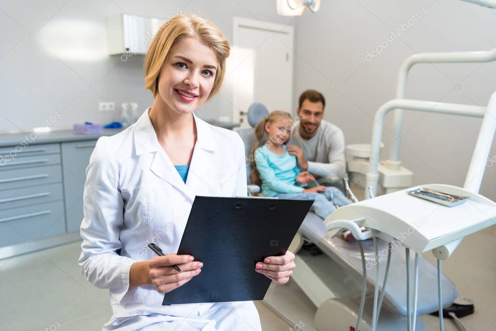 beautiful female dentist with clipboard looking at camera while little child and her father sitting on background