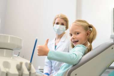 beautiful little child showing thumb up while sitting in chair at dentist office clipart
