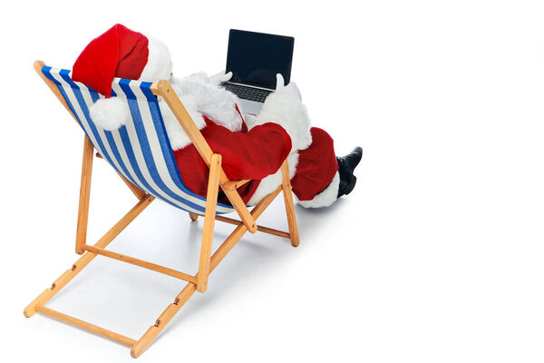 back view of santa claus using laptop with blank screen while relaxing on beach chair isolated on white