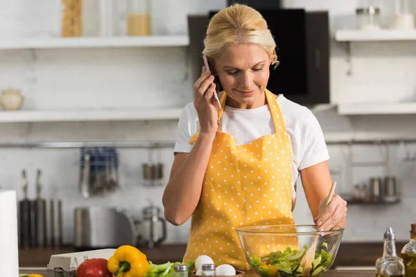 Sorrindo Mulher Madura Avental Falando Por Smartphone Enquanto Cozinha Salada — Fotos gratuitas