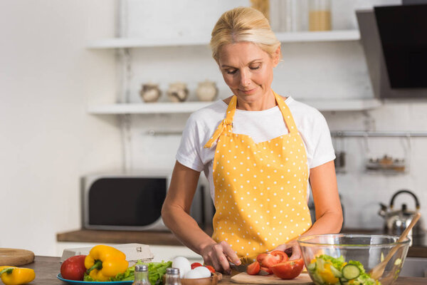 beautiful mature woman in apron cooking vegetable salad in kitchen   