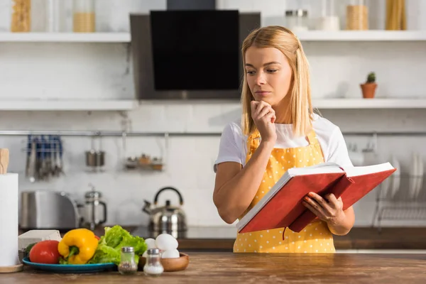 Mujer Joven Reflexiva Delantal Sosteniendo Libro Cocina Mirando Los Ingredientes —  Fotos de Stock