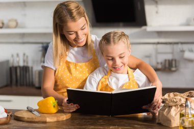 happy mother and daughter reading cookbook while cooking together clipart