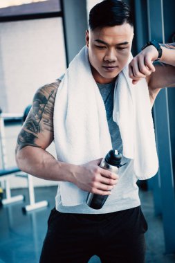 young tired asian sportsman with towel and bottle of water after long workout clipart