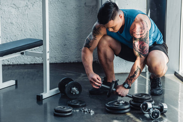 tattooed sportsman assembling dumbbells at gym