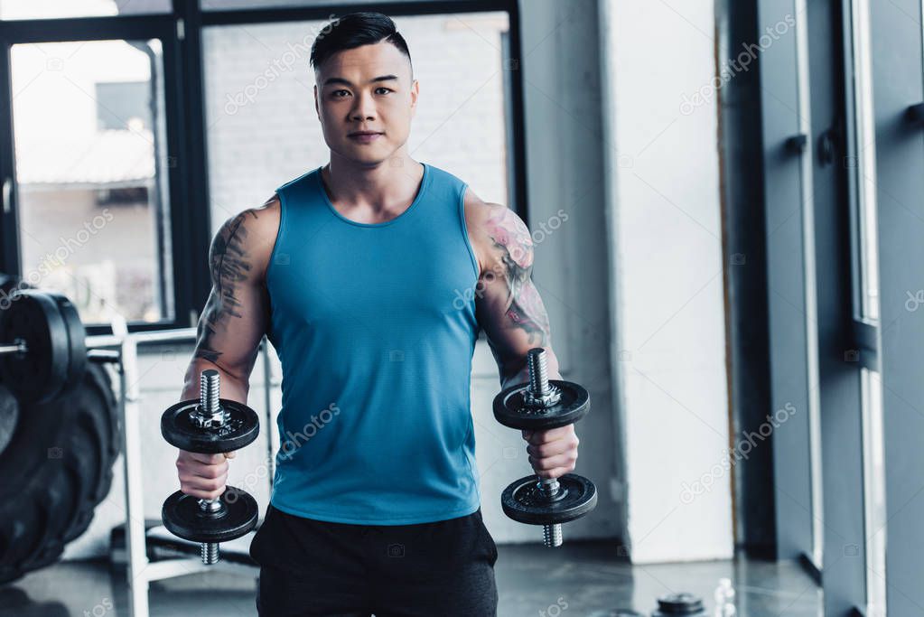 concentrated young asian sportsman exercising with dumbbells in gym