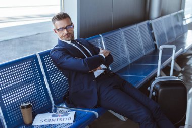 businessman in glasses sitting near window with newspaper and paper cup at departure lounge  clipart