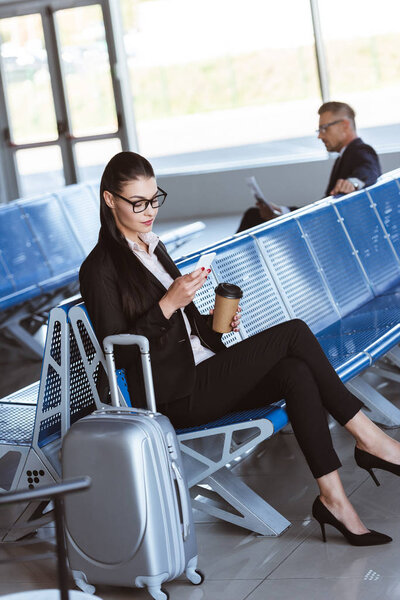 young businesswoman in glasses with baggage using smartphone at airport