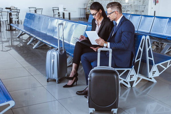 молодая бизнесвумен, пишущая в блокноте, в то время как бизнесмен использует цифровой планшет в аэропорту
