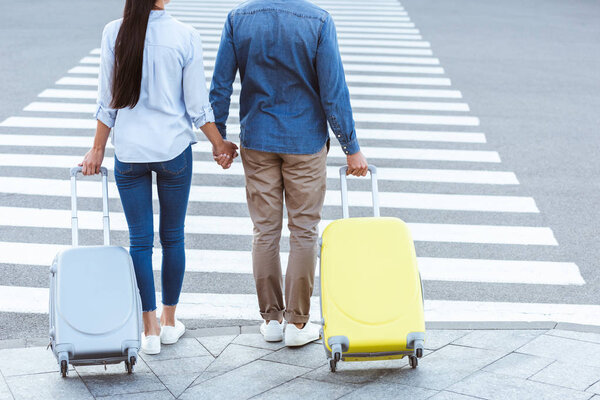 A cropped view of a couple of travelers crossing pedestrian, holding hands and pulling their luggage