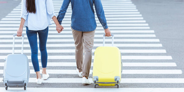 обрезанный вид на пару туристов, пересекающих пешеходную зону, держащихся за руки и вытаскивающих свой багаж
