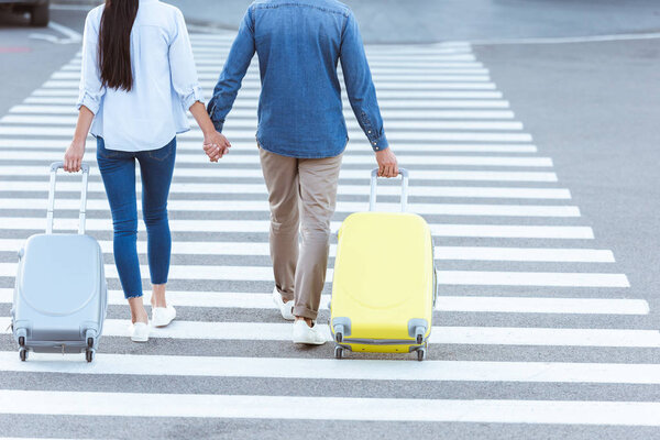 Пара туристов, переходящих пешеходов, держащихся за руки и тащащих свой багаж
