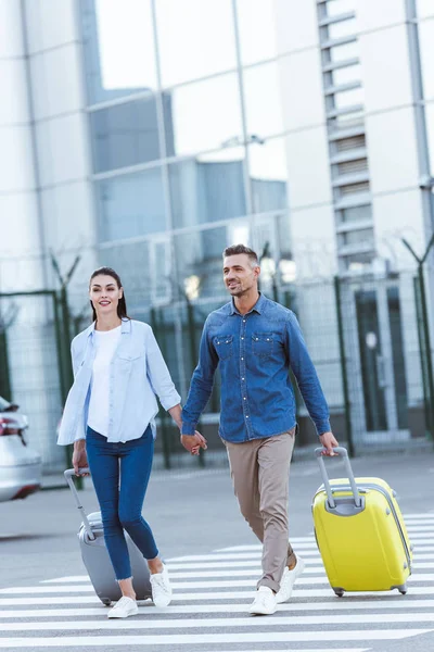 Smiling Couple Tourists Crossing Pedestrian Holding Hands Pulling Luggage — Free Stock Photo