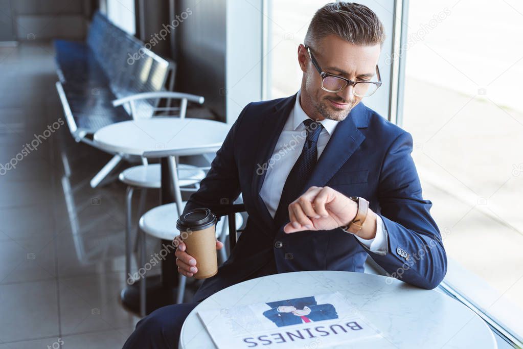 handsome businessman in glasses holding coffee to go and looking at watches on hand