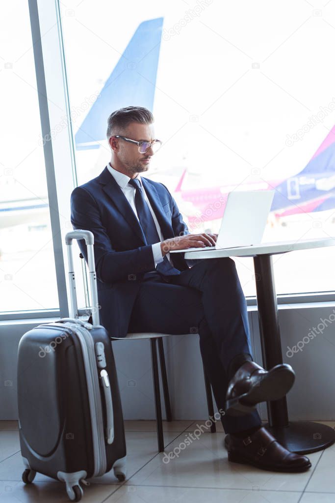 adult businessman in glasses typing on laptop at table in airport