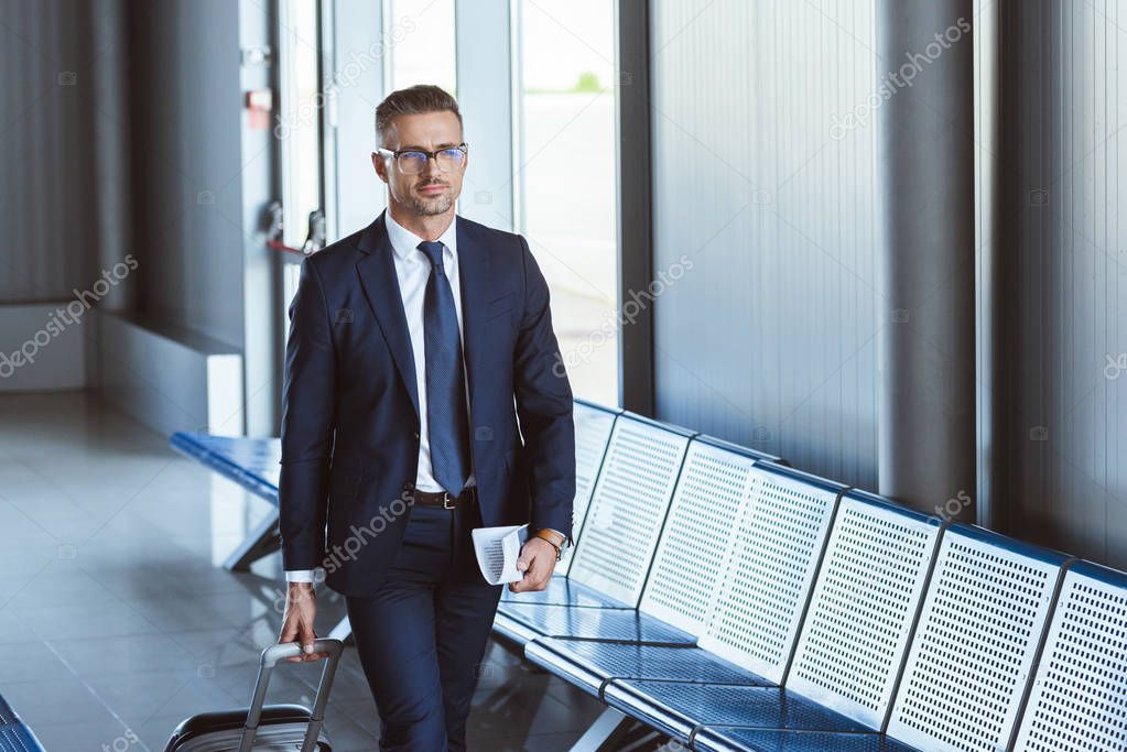 adult handsome businessman in glasses with baggage and tickets walking at airport