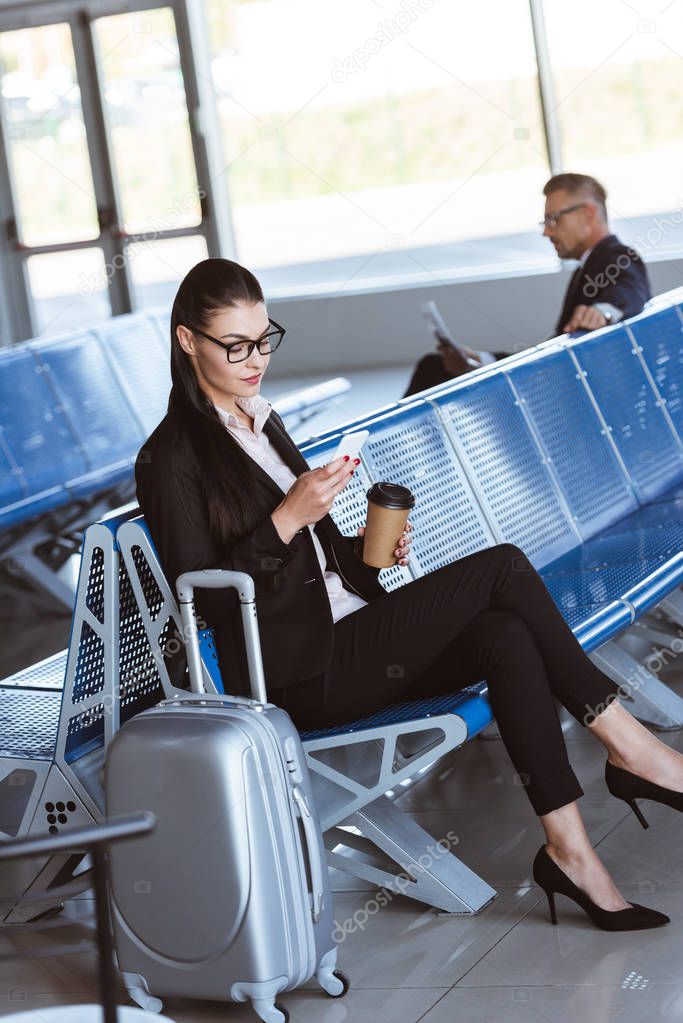 young businesswoman in glasses with baggage using smartphone at airport