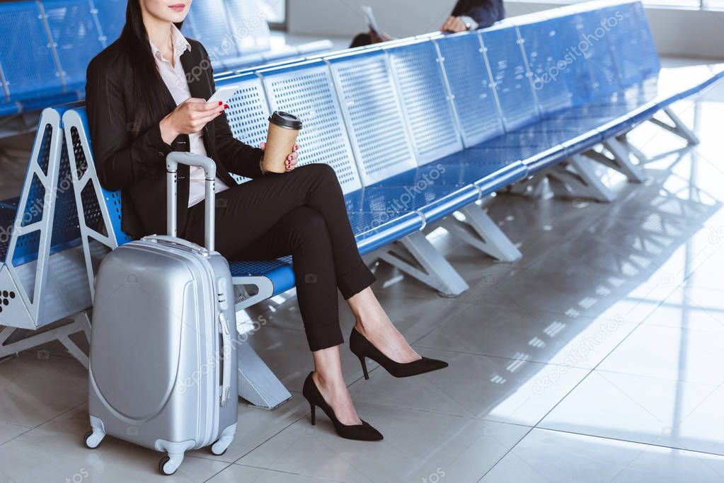 businesswoman with coffee to go using smartphone in departure lounge at airport