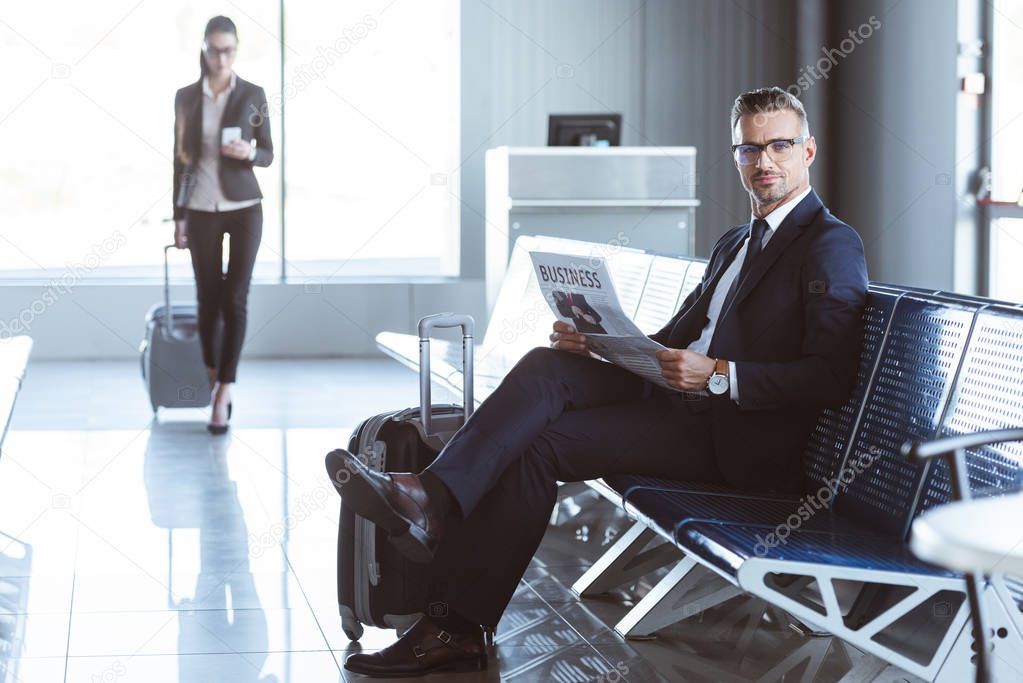adult businessman reading newspaper while businesswoman walking with luggage at departure lounge at airport