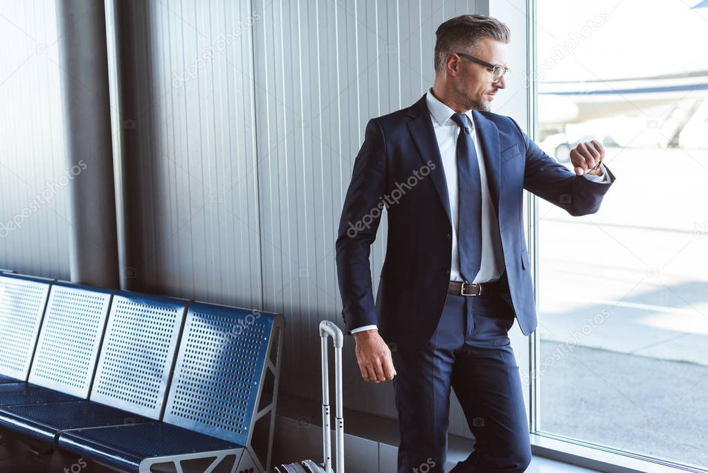 businessman standing at departure lounge near window and looking at watch in airport