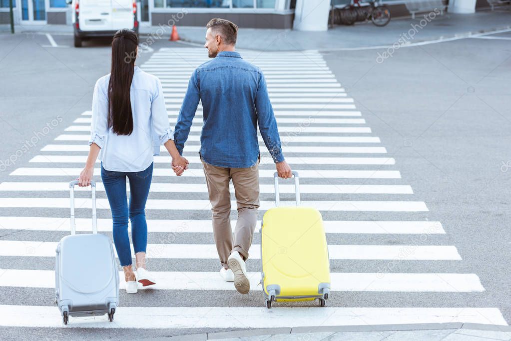 A couple of tourists crossing pedestrian, holding hands and pulling their luggage