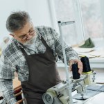 Focused middle aged tailor in apron setting strings on sewing machine at workshop