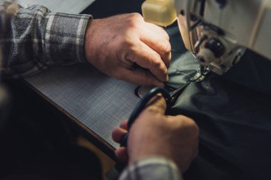 cropped image of male handbag craftsman cutting leather near sewing machine at studio clipart