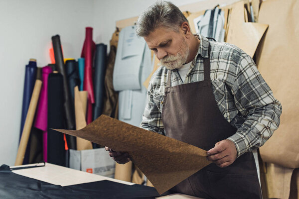 middle aged tailor in apron looking at cardboard in workshop 