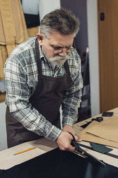 middle aged handbag craftsman in apron and eyeglasses cutting leather by scissors at workshop