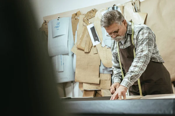 selective focus of mature handbag craftsman in apron making measurements on leather at workshop