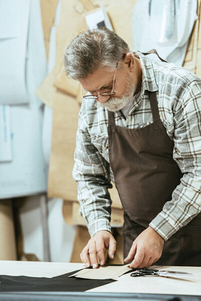 serious middle aged handbag craftsman in apron and eyeglasses working at studio