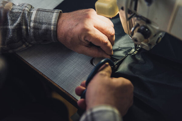cropped image of male handbag craftsman cutting leather near sewing machine at studio