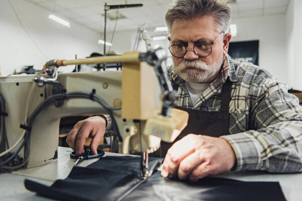 male tailor in apron and eyeglasses working on sewing machine at studio
