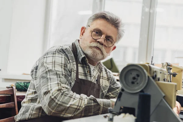 Portrait Mature Male Tailor Apron Eyeglasses Sitting Sewing Machine Studio — Free Stock Photo