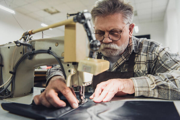 selective focus of mature male tailor in apron and eyeglasses working on sewing machine at studio