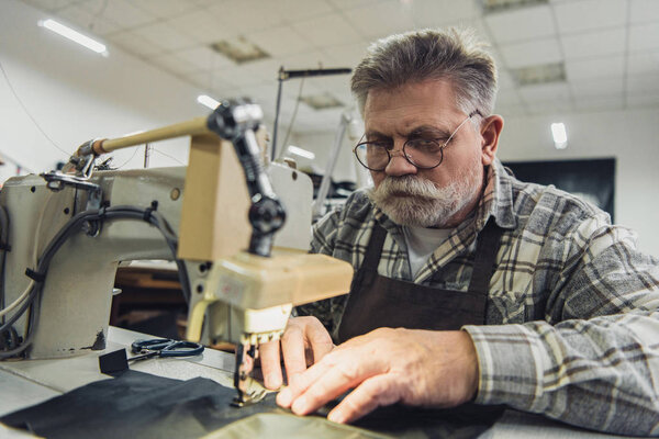 concentrated mature male tailor in apron and eyeglasses working on sewing machine at studio