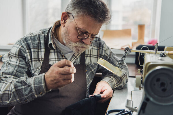 selective focus of middle aged male tailor in eyeglasses and apron sewing at workshop