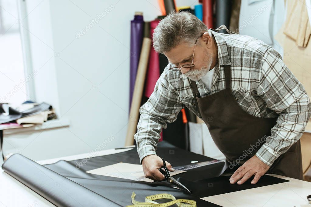 male craftsman in apron and eyeglasses cutting leather by scissors at workshop