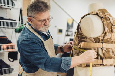 mature male tailor making measurements on military vest at workshop clipart