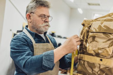 selective focus of middle aged male tailor making measurements on military vest at workshop clipart